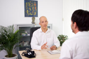 Arzt und Patient sitzen sich in einer Praxis gegenüber und führen ein Diagnose-Gespräch über Hämorrhoiden.