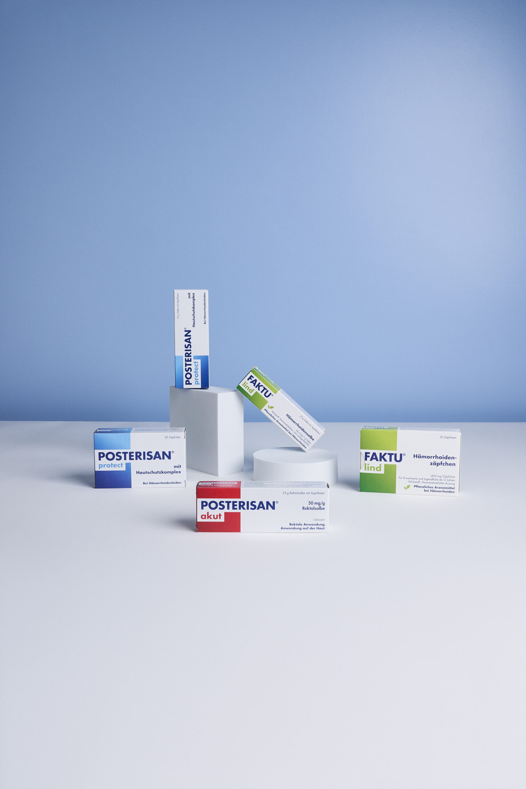 Arrangierte Produktdarstellung mit Posterisan akut, Posterisan protect Salbe und Zäpfchen sowie FAKTU lind Salbe und Zäpfchen auf einer weißen Fläche vor blauem Hintergrund.